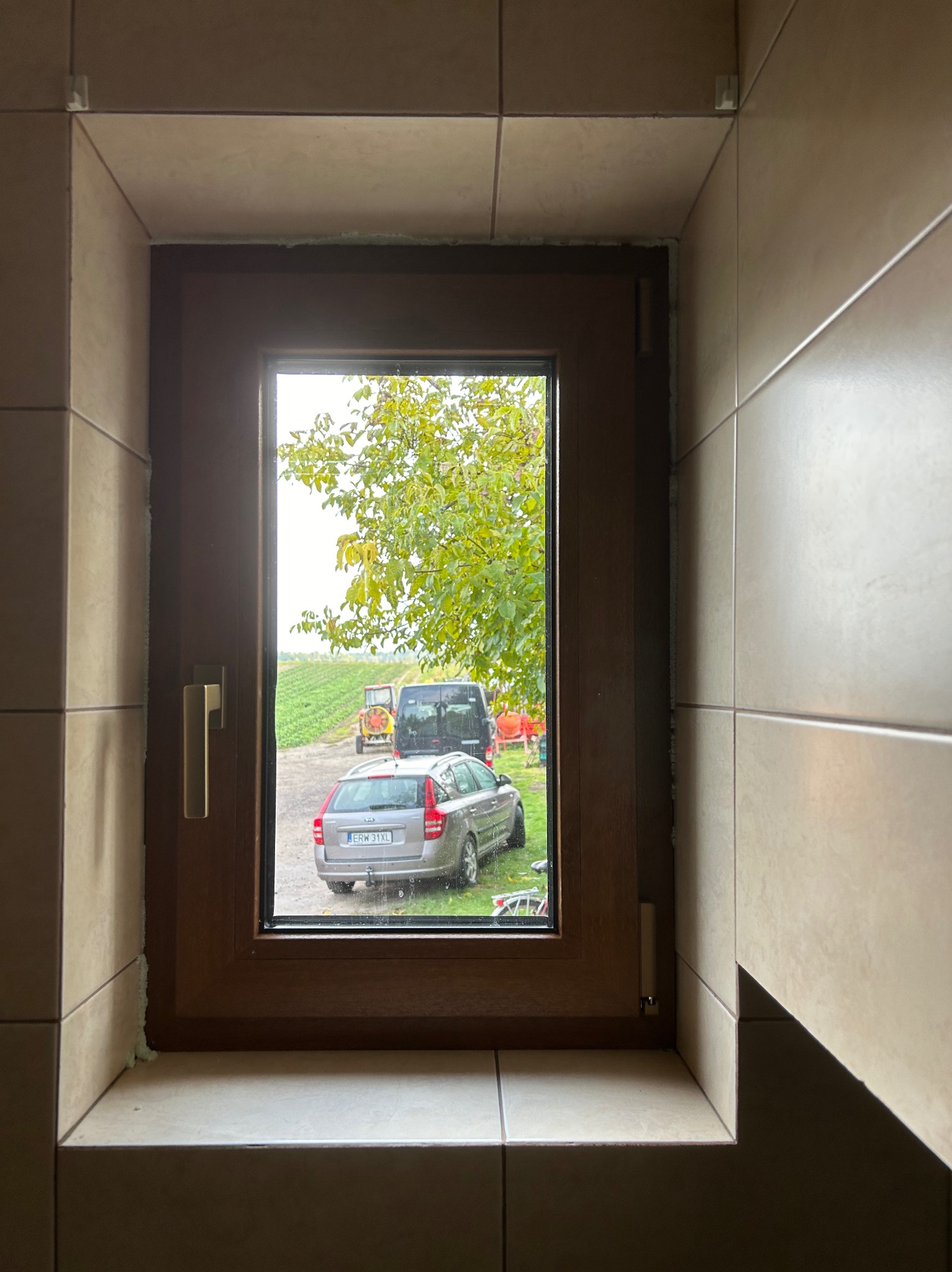 Brązowe okno z widokiem na pole z traktorem i samochodem, widoczne ślady pianki montażowej przy ramie, wnętrze wyłożone beżowymi płytkami.