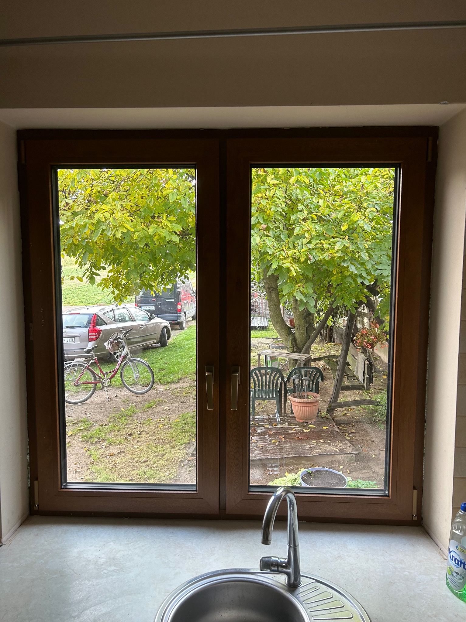 Dwuskrzydłowe okno z brązowymi ramami, widok na podwórze z rowerem, samochodami i drzewem, zlewozmywak w dolnej części kadru.