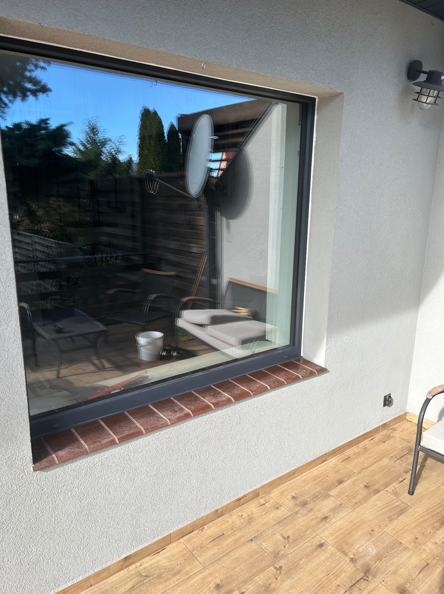 Nowe okno z ciemną ramą i ceglanym parapetem zewnętrznym, widok z tarasu na ogród odbity w szybie.