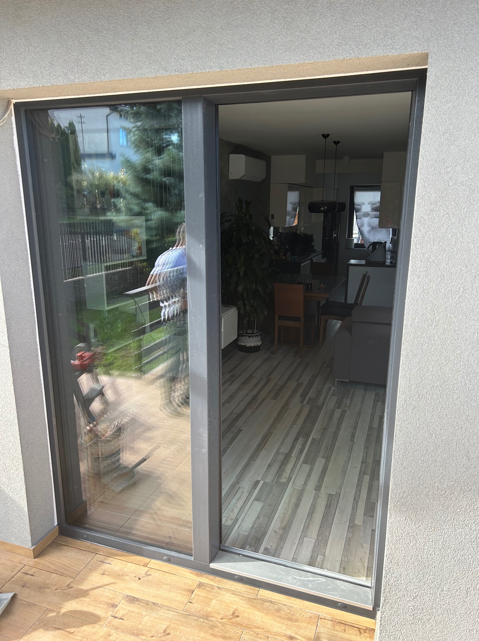 Nowoczesne, szare drzwi przesuwne z widokiem na wnętrze domu z drewnianą podłogą i kuchnią w tle oraz na zewnątrz z widocznym ogrodem i narzędziami malarskimi.