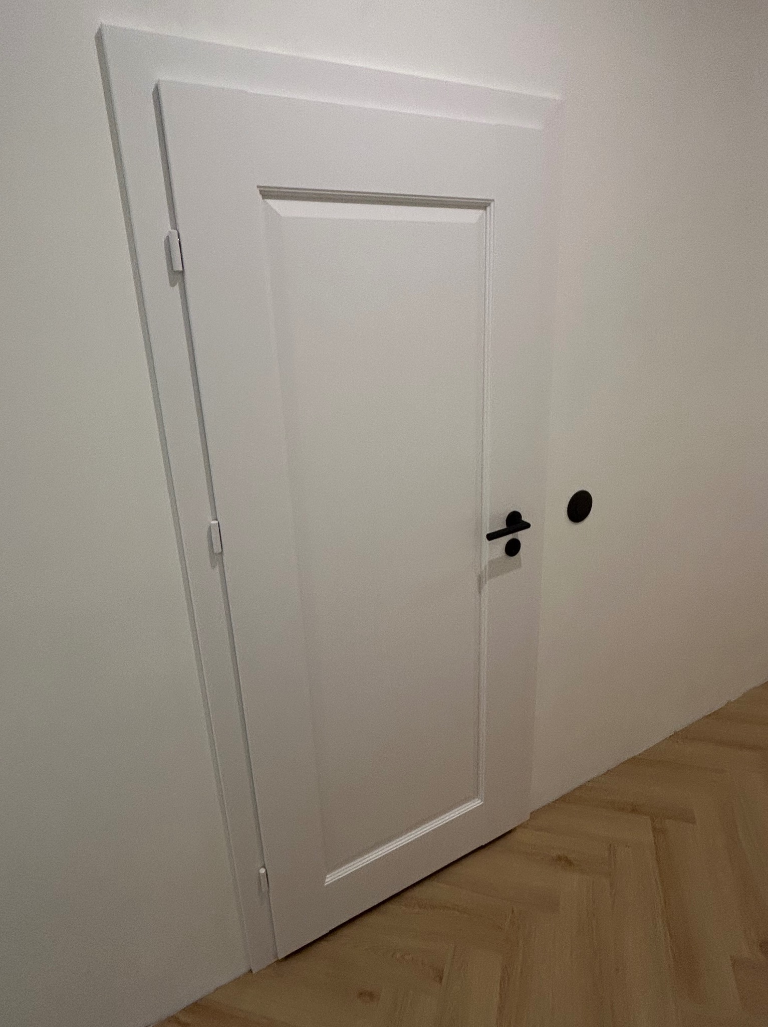 Białe drzwi wewnętrzne z czarną klamką i minimalistyczną ościeżnicą, widoczne na tle jasnej ściany i parkietu w jodełkę.
