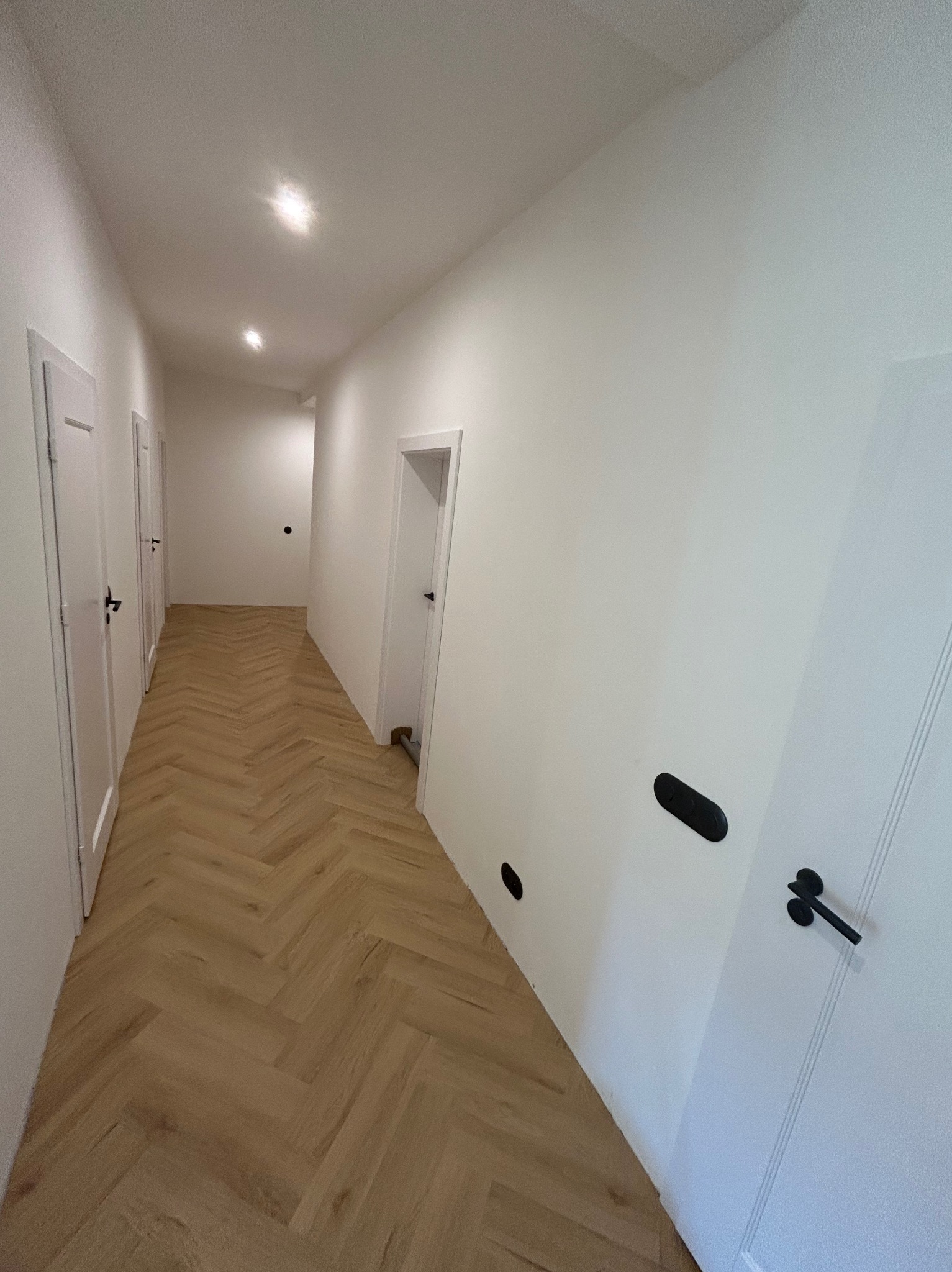 Korytarz z jasną podłogą ułożoną we wzór jodełki, białymi ścianami i białymi drzwiami z czarnymi klamkami; widoczne czarne kontakty na ścianie.