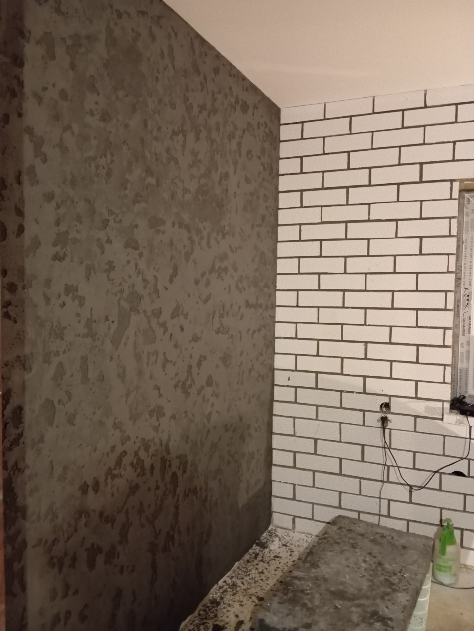 Ściana z dekoracyjną, szarą fakturą przypominającą beton, sąsiadująca z białą ścianą z cegły, widoczne gniazdko elektryczne i butelka z rozpylaczem.