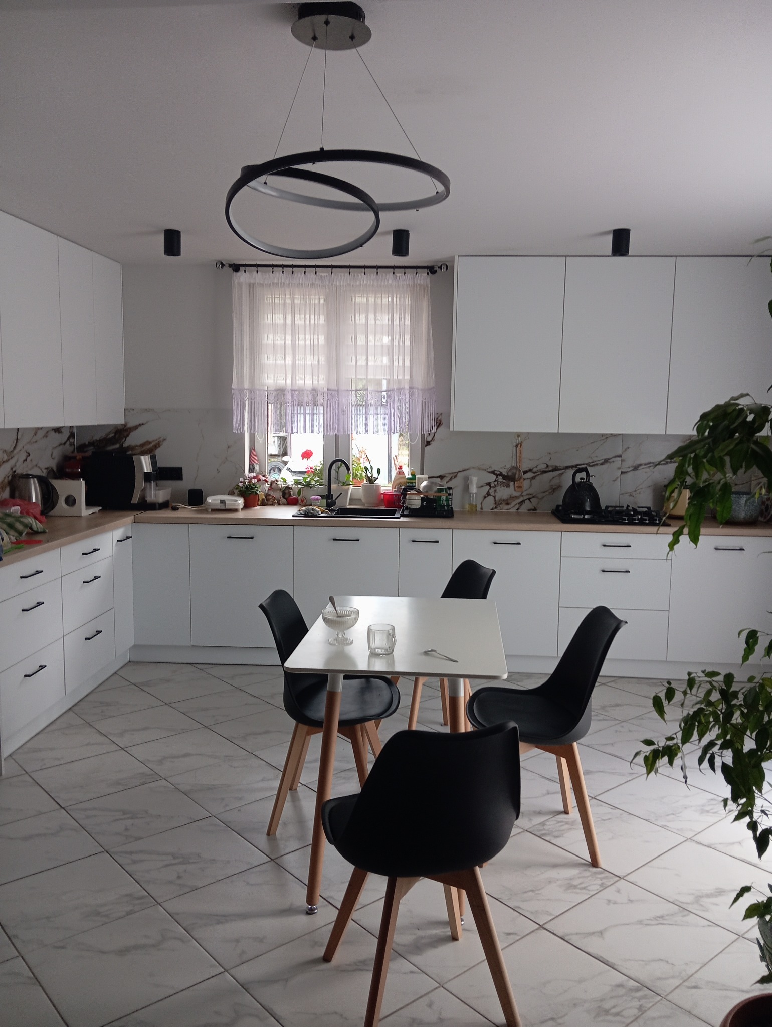 Jasna kuchnia z białymi szafkami, marmurowym blatem i podłogą, okrągłym czarnym żyrandolem, stołem z czterema czarnymi krzesłami i rośliną w doniczce.