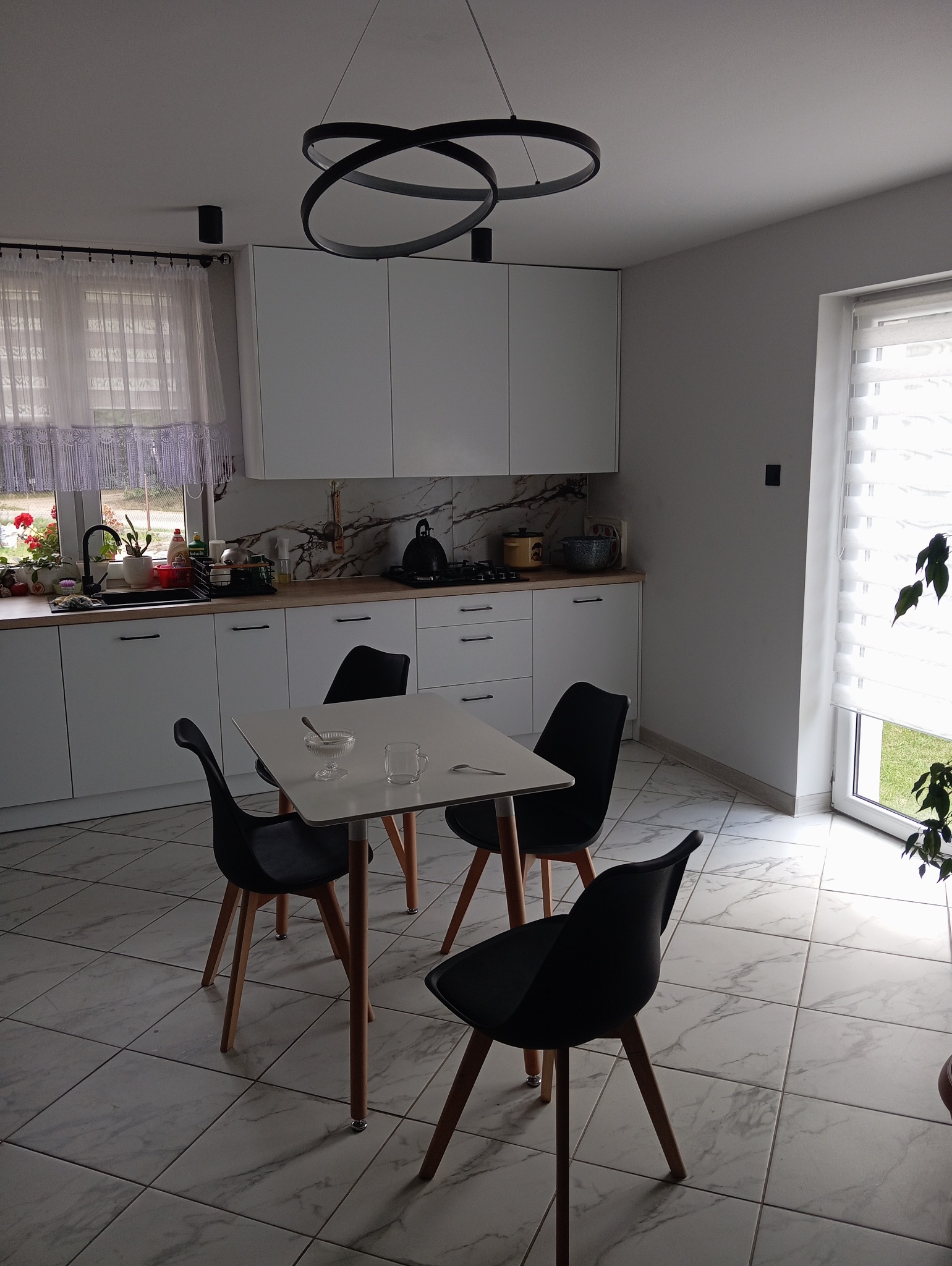 Nowoczesna, jasna kuchnia z białymi szafkami, marmurowym panelem nad blatem i designerską lampą sufitową nad kwadratowym stołem z czarnymi krzesłami.