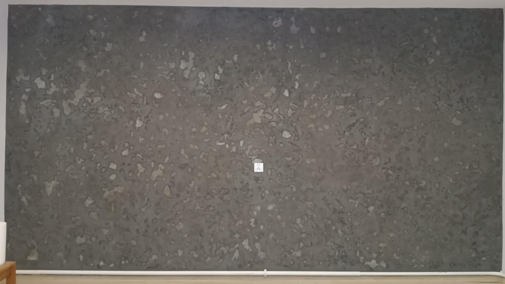 Dekoracyjna ściana w odcieniach szarości z efektem nieregularnych plam, widoczny kontakt elektryczny na środku ściany, listwa przypodłogowa wzdłuż dolnej krawędzi.