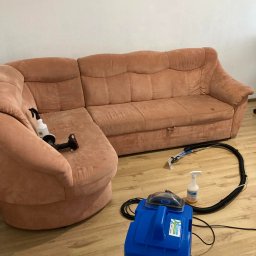 Władysław Kudriaszow BigMax Clean - Różowa, narożna kanapa z widocznymi plamami, obok niej niebieski odkurzacz piorący z przezroczystą końcówką i przewodem oraz butelka ze środkiem czyszczącym.