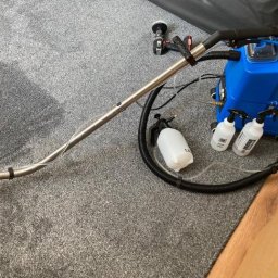 Władysław Kudriaszow BigMax Clean - Profesjonalny sprzęt do czyszczenia dywanów: niebieski odkurzacz ekstrakcyjny, dysza ssąca na długiej rurze, butelki ze środkiem czyszczącym i dodatkowa szczotka na szarym dywanie.