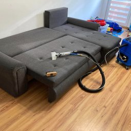 Władysław Kudriaszow BigMax Clean - Rozłożona szara kanapa narożna z funkcją spania, czyszczona za pomocą niebieskiego odkurzacza piorącego i małej szczotki. W tle zabawki dziecięce.