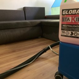 Władysław Kudriaszow BigMax Clean - Ciemnoszara narożna kanapa widoczna w tle, na pierwszym planie niebieski odkurzacz piorący z podłączonym wężem i pojemnikiem z napisem 'Neutralizator' na podłodze z paneli.