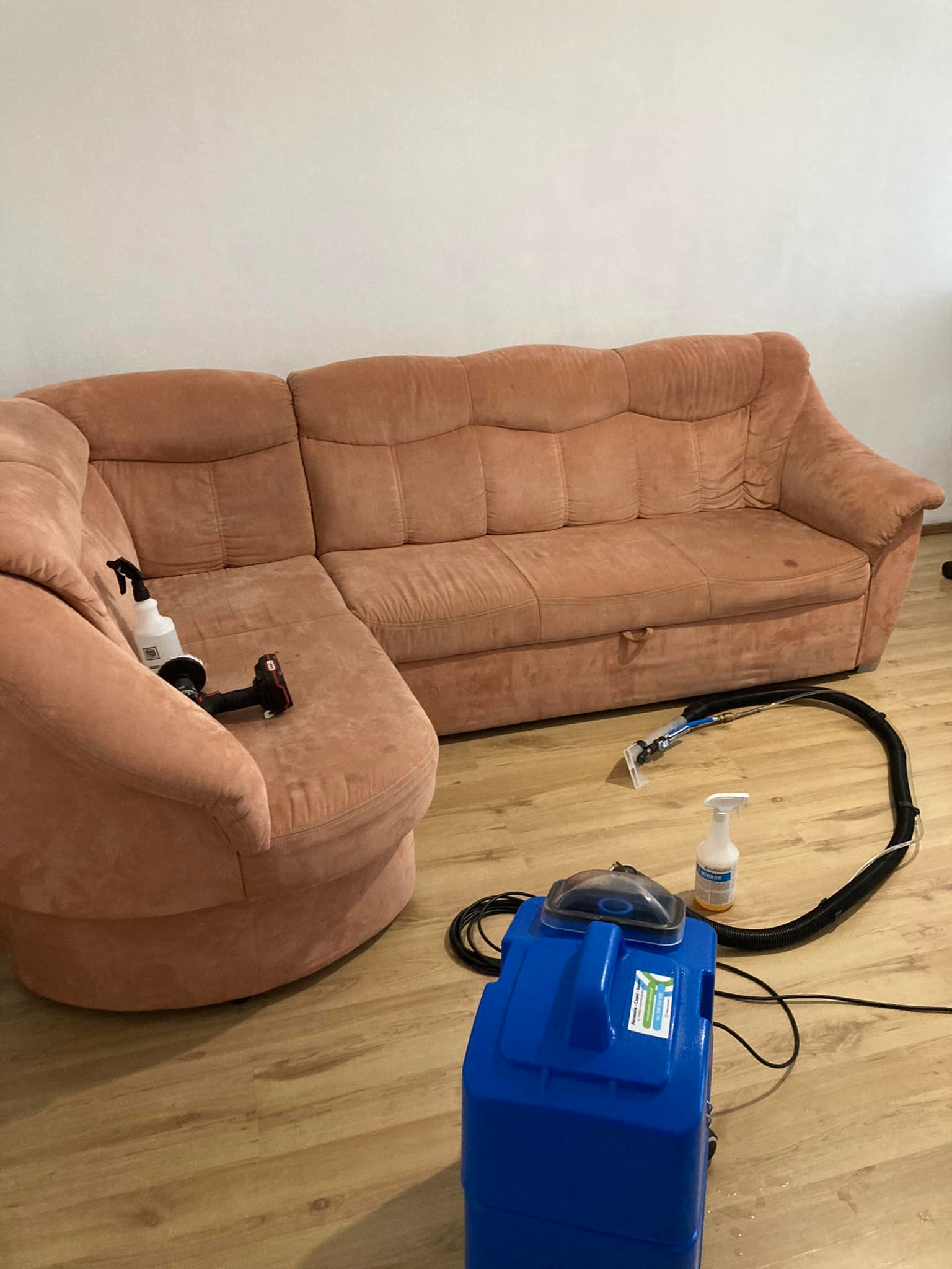 Różowa, narożna kanapa z widocznymi plamami, obok niej niebieski odkurzacz piorący z przezroczystą końcówką i przewodem oraz butelka ze środkiem czyszczącym.