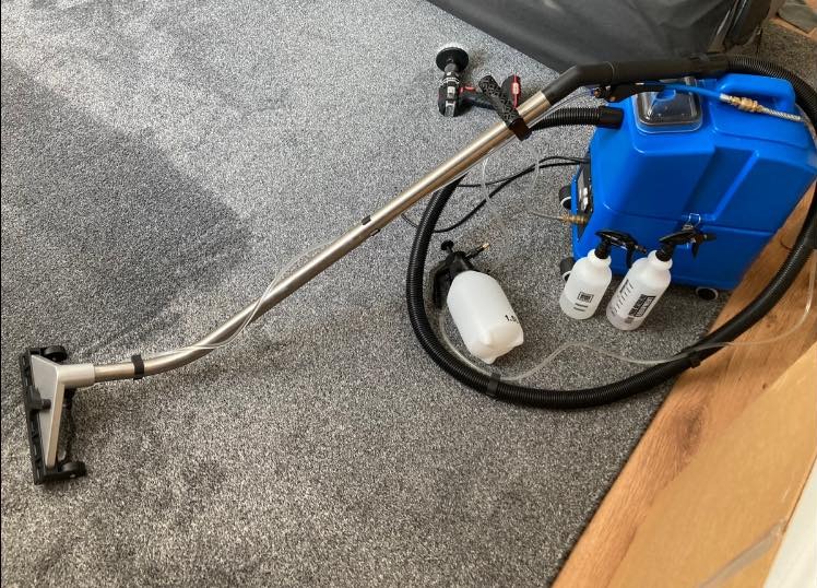 Profesjonalny sprzęt do czyszczenia dywanów: niebieski odkurzacz ekstrakcyjny, dysza ssąca na długiej rurze, butelki ze środkiem czyszczącym i dodatkowa szczotka na szarym dywanie.