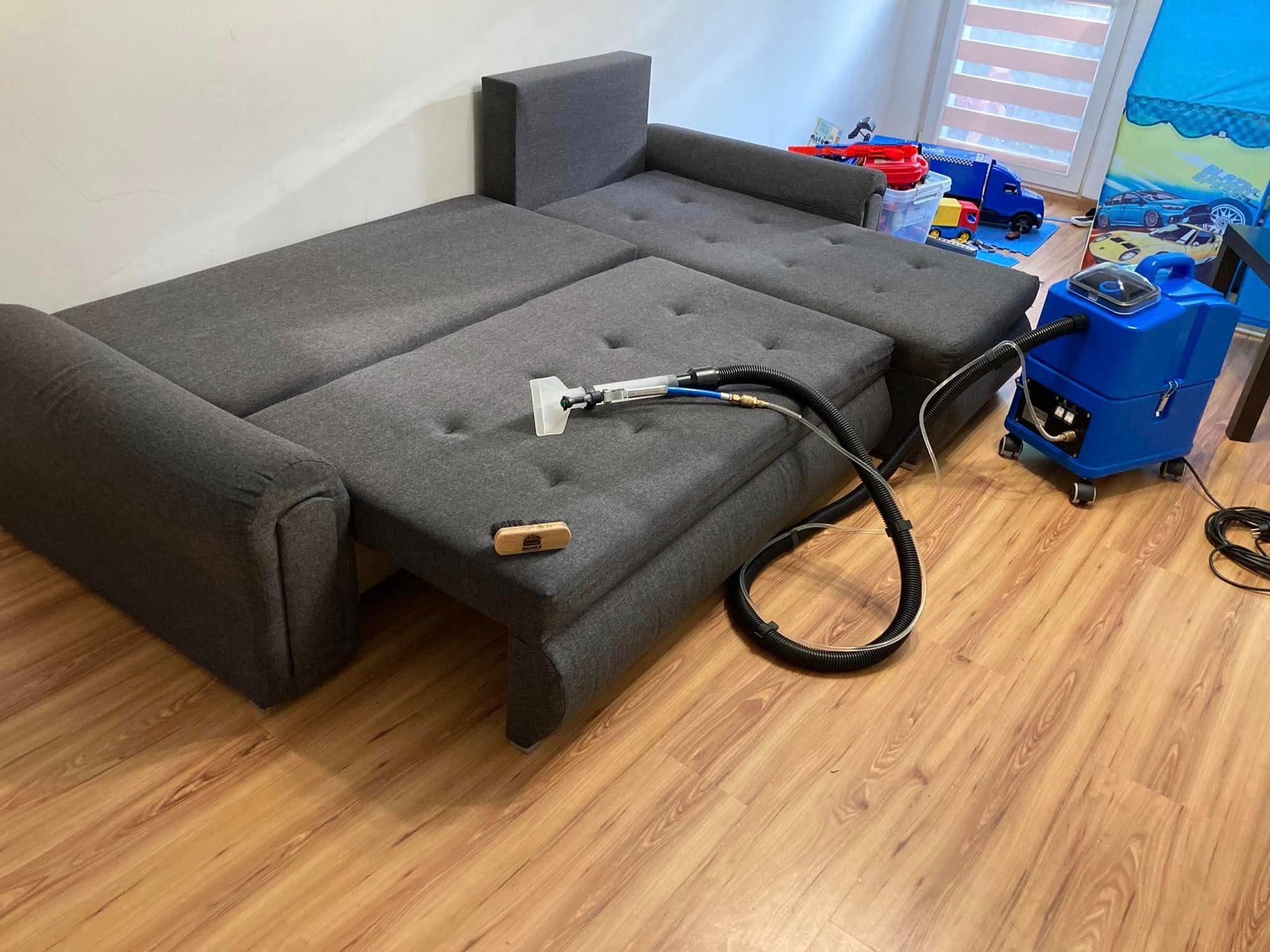 Rozłożona szara kanapa narożna z funkcją spania, czyszczona za pomocą niebieskiego odkurzacza piorącego i małej szczotki. W tle zabawki dziecięce.