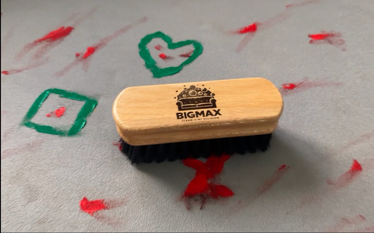 Drewniana szczotka z czarnym włosiem i logo firmy BIGMAX na jasnoszarym dywanie z czerwonymi i zielonymi plamami farby w kształcie serca, kwadratu i innych wzorów.