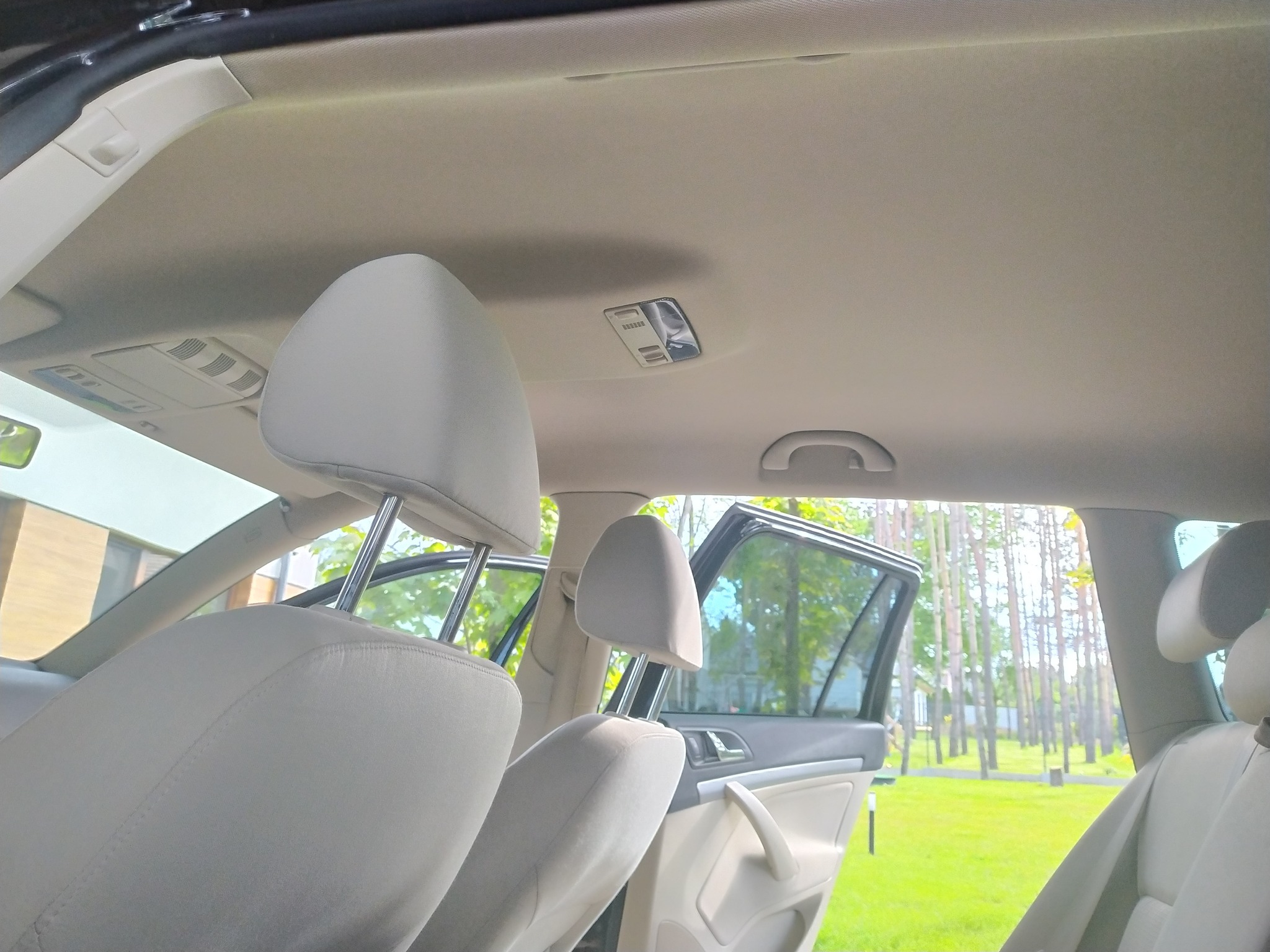 Wnętrze samochodu z jasną tapicerką, widoczne fotele z zagłówkami, podsufitka i otwarte drzwi, przez które widać zieleń na zewnątrz.