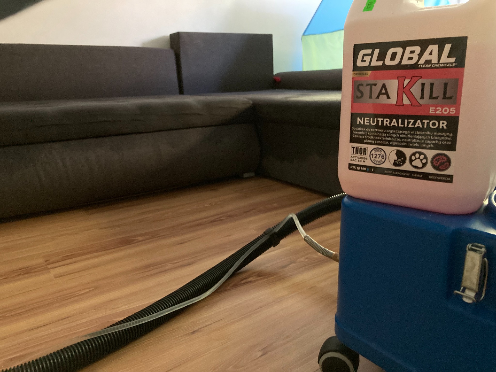 Ciemnoszara narożna kanapa widoczna w tle, na pierwszym planie niebieski odkurzacz piorący z podłączonym wężem i pojemnikiem z napisem 'Neutralizator' na podłodze z paneli.