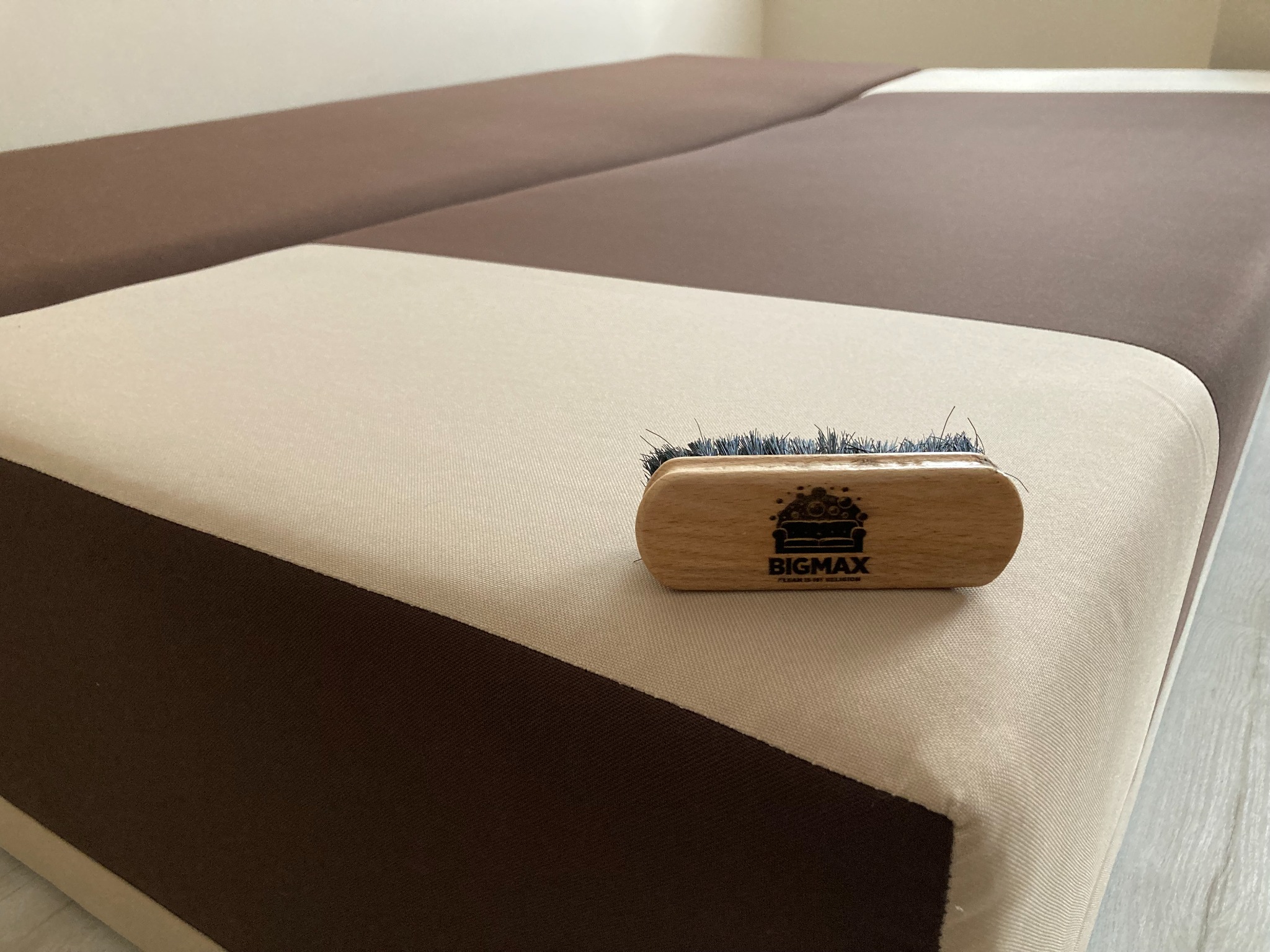 Szczotka do czyszczenia tapicerki z logo firmy BIGMAX leżąca na dwukolorowej kanapie w odcieniach beżu i brązu.