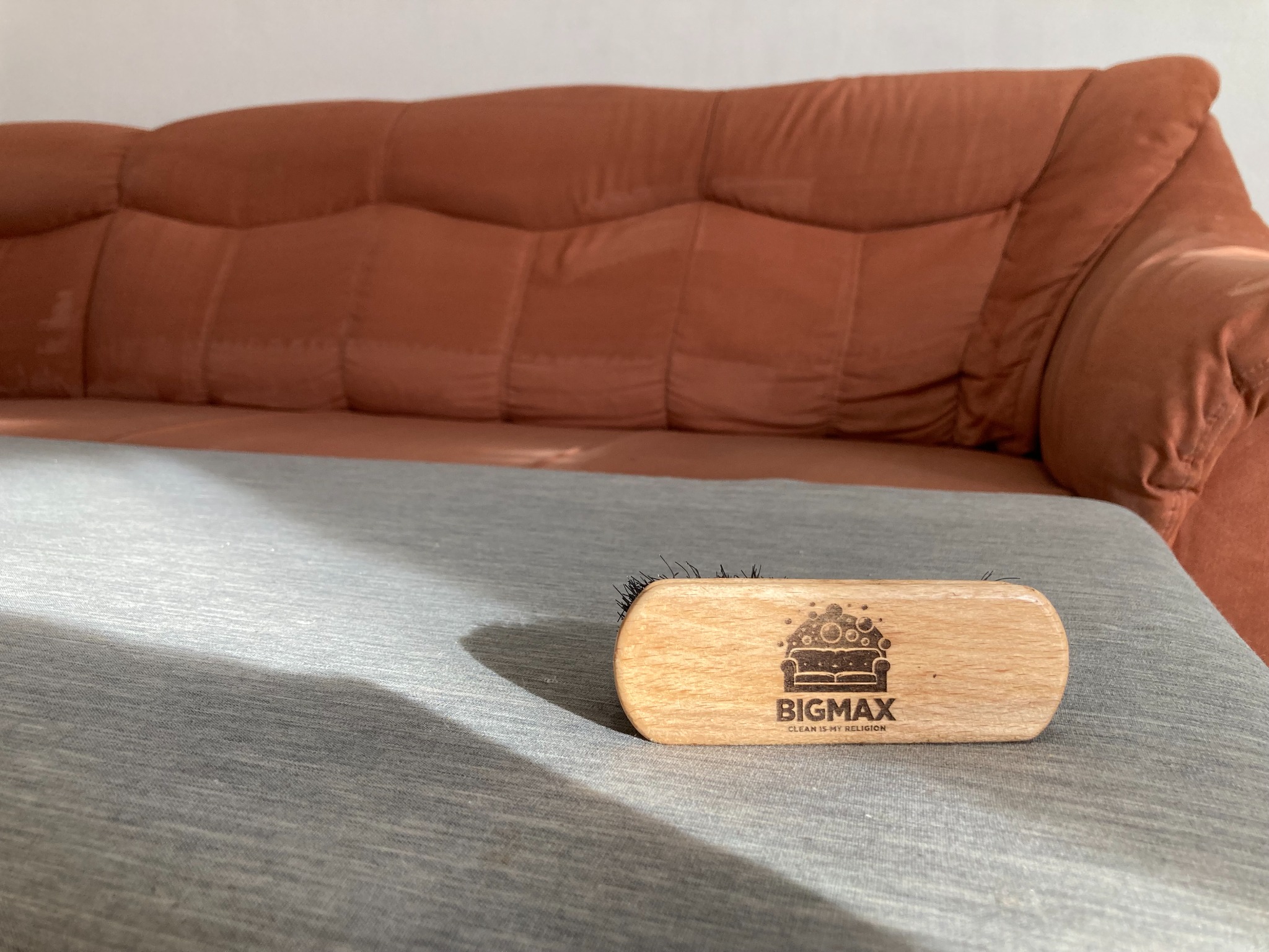 Szczotka z logo firmy 'BIGMAX Clean is my religion' leży na szarym materiale sofy, w tle widoczna część pomarańczowej kanapy.