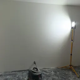 Świeżo pomalowana ściana w pokoju, widoczny brudny od gładzi odkurzacz przemysłowy Bosch, lampa na statywie oświetlająca ścianę, podłoga zabezpieczona folią malarską.