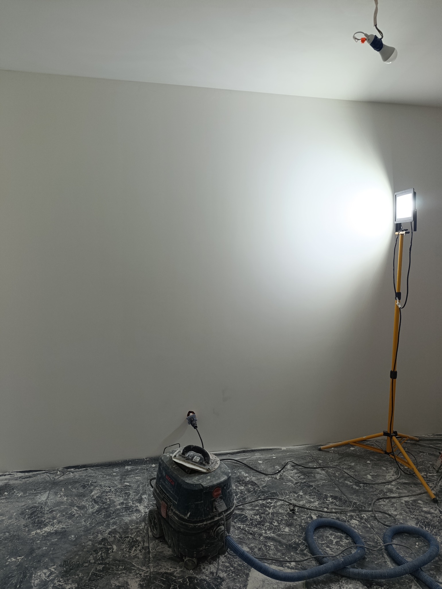 Świeżo pomalowana ściana w pokoju, widoczny brudny od gładzi odkurzacz przemysłowy Bosch, lampa na statywie oświetlająca ścianę, podłoga zabezpieczona folią malarską.