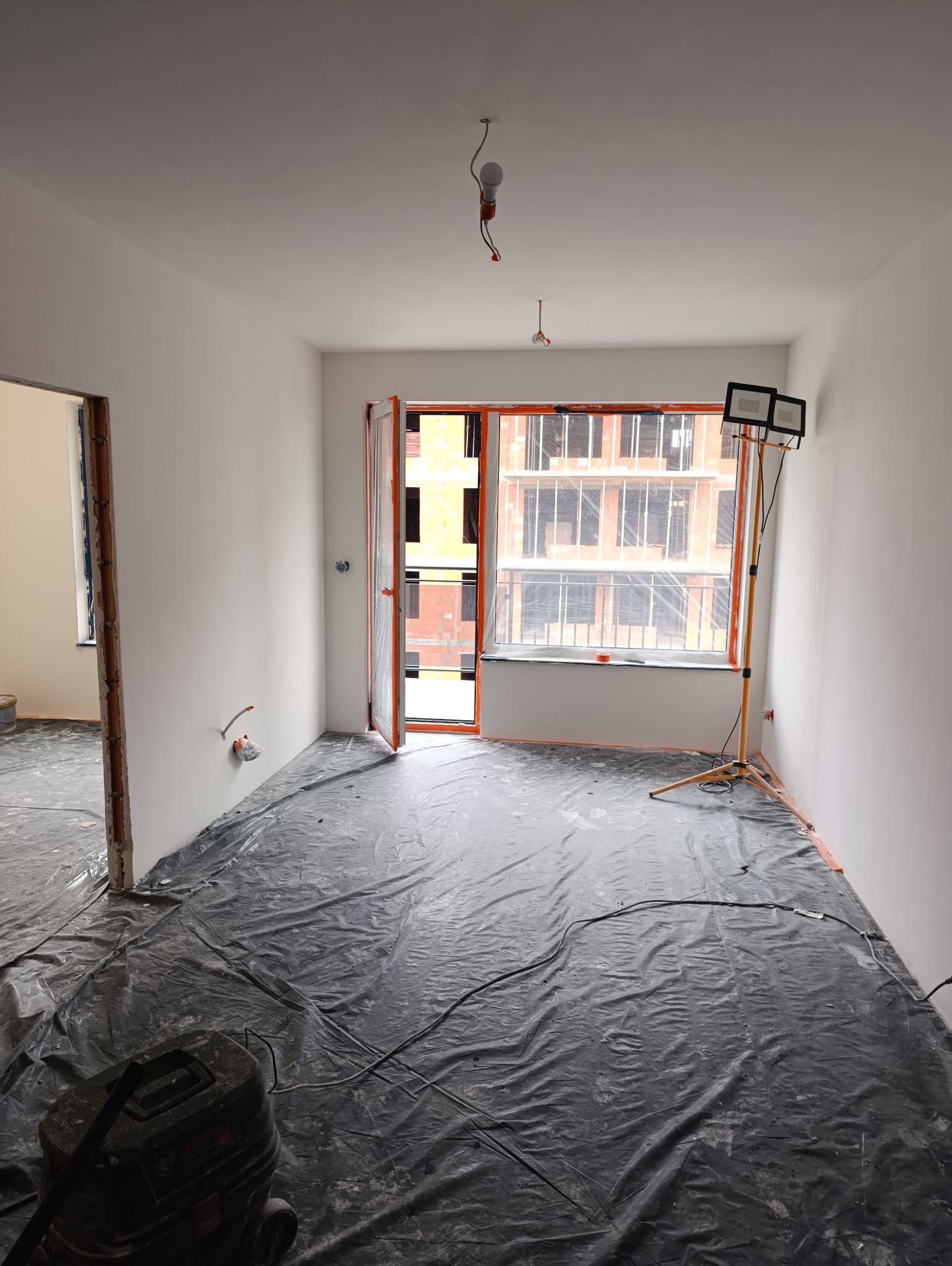 Pokój w trakcie odświeżania: świeżo pomalowane ściany na biało, podłoga zabezpieczona czarną folią, widoczny odkurzacz budowlany i lampa halogenowa na statywie przy oknie.