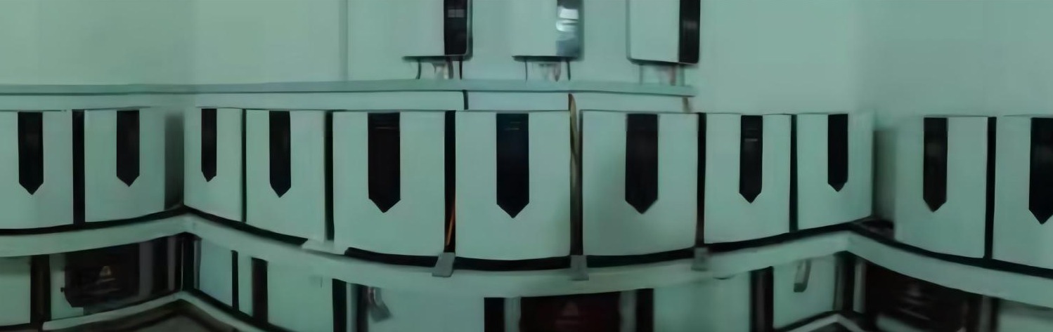 Bateria modułowa z białymi obudowami i czarnymi paskami, ustawiona na półkach w pomieszczeniu o jasnych ścianach.