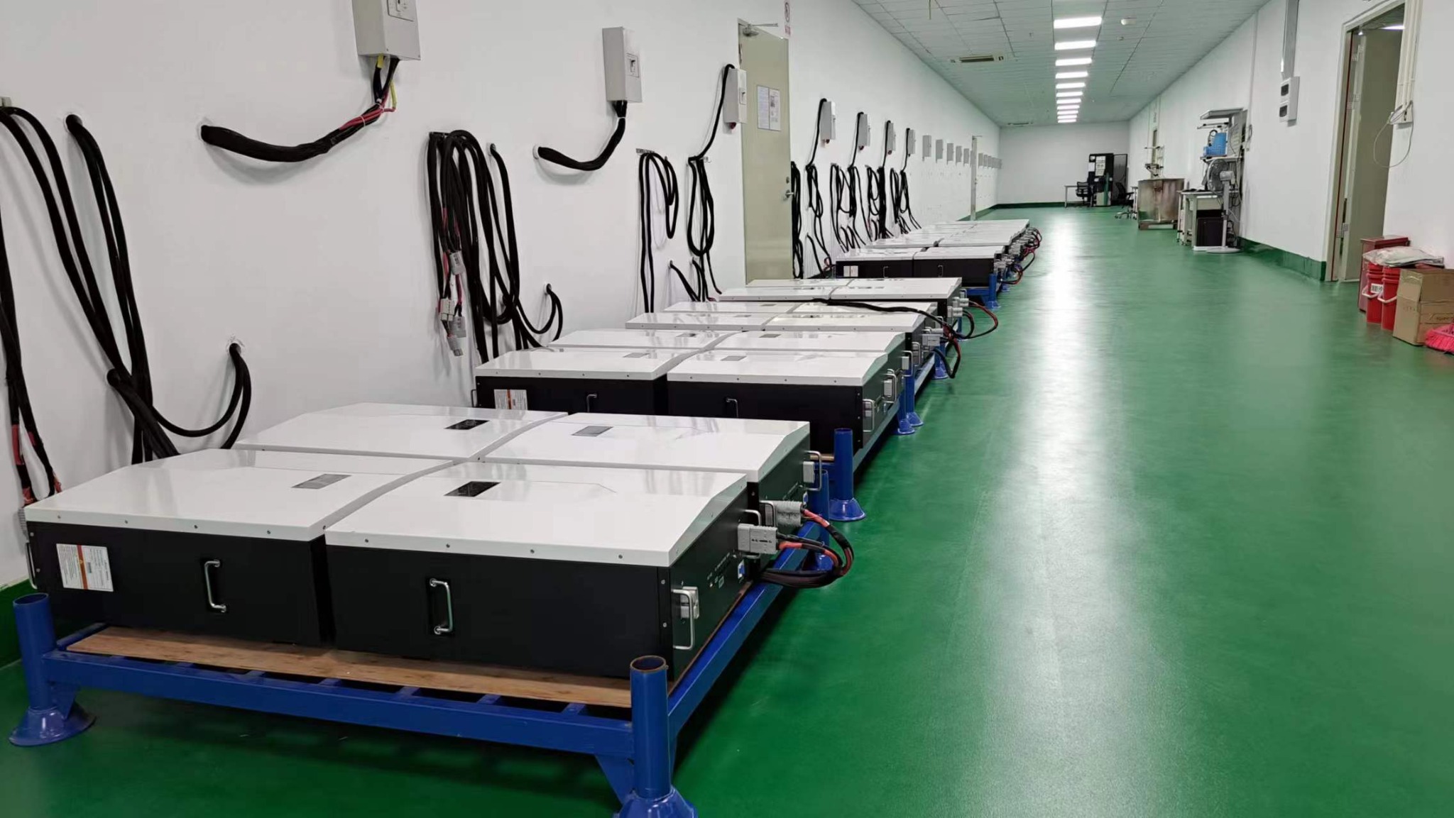 Rząd akumulatorowych systemów magazynowania energii na niebieskich paletach w hali w okolicach Wrocławia, z przewodami biegnącymi do gniazd na ścianie.