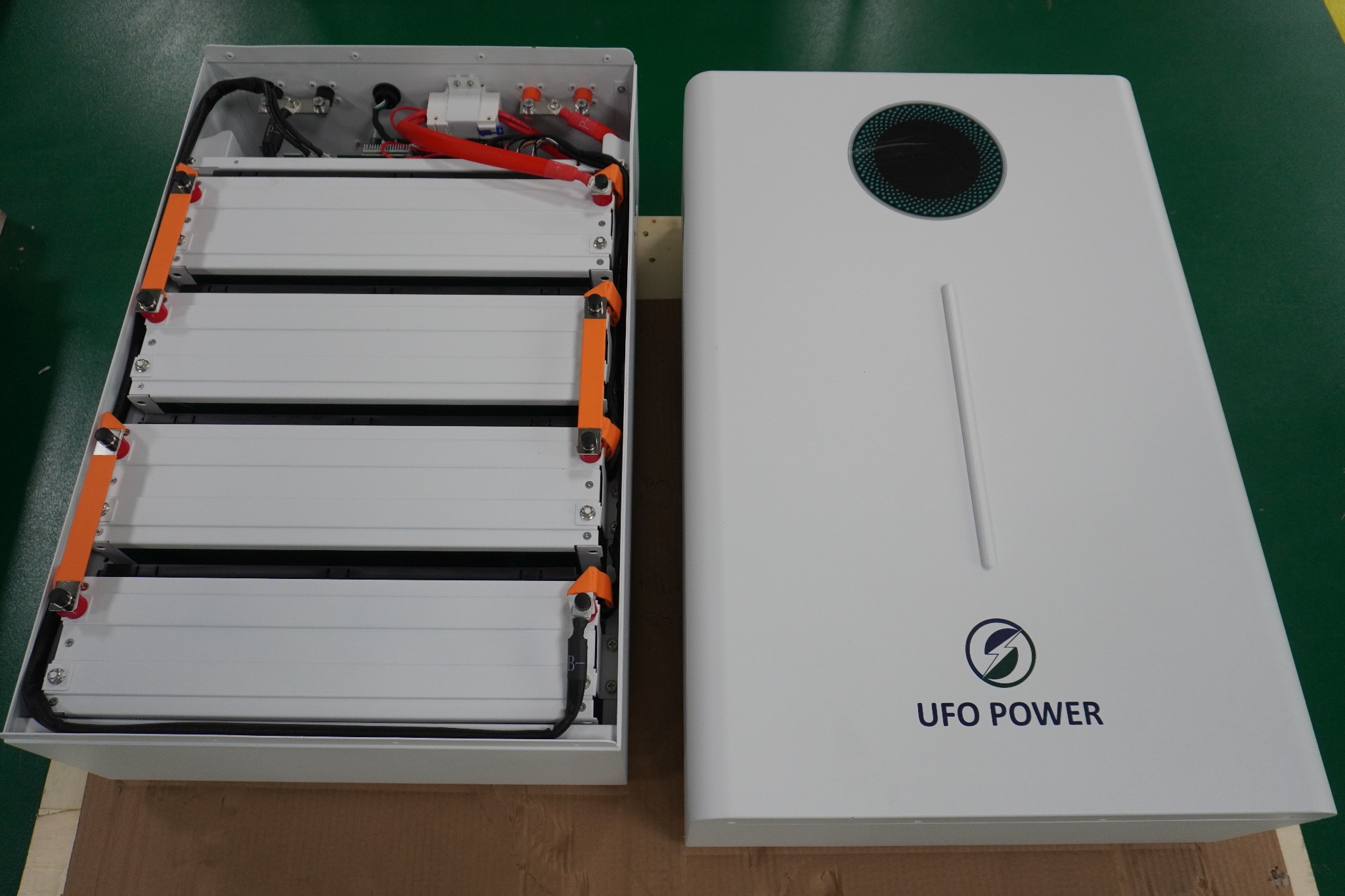 Otwarta obudowa akumulatorowa z czterema białymi modułami bateryjnymi i zamknięta obudowa z logo UFO Power na tle zielonego blatu.