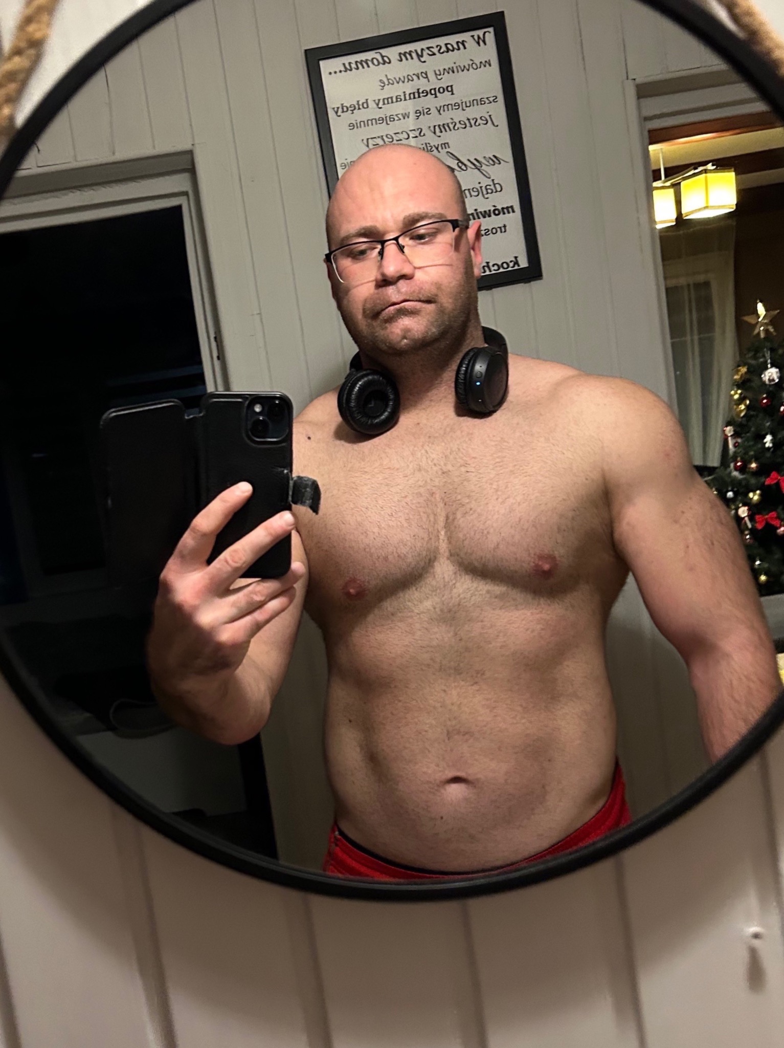 Mężczyzna w okularach i słuchawkach na szyi, robiący selfie w lustrze, prezentujący swoją umięśnioną sylwetkę, w tle widoczna choinka.