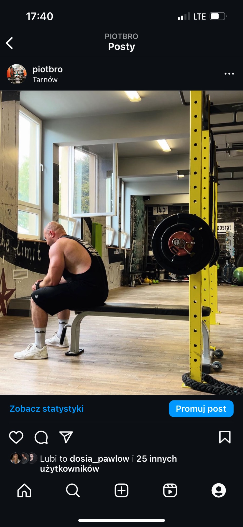 Mężczyzna w czarnym tank topie siedzi na ławce w siłowni, obok żółtej klatki do ćwiczeń ze sztangą, w tle widoczne graffiti na ścianie i okno.