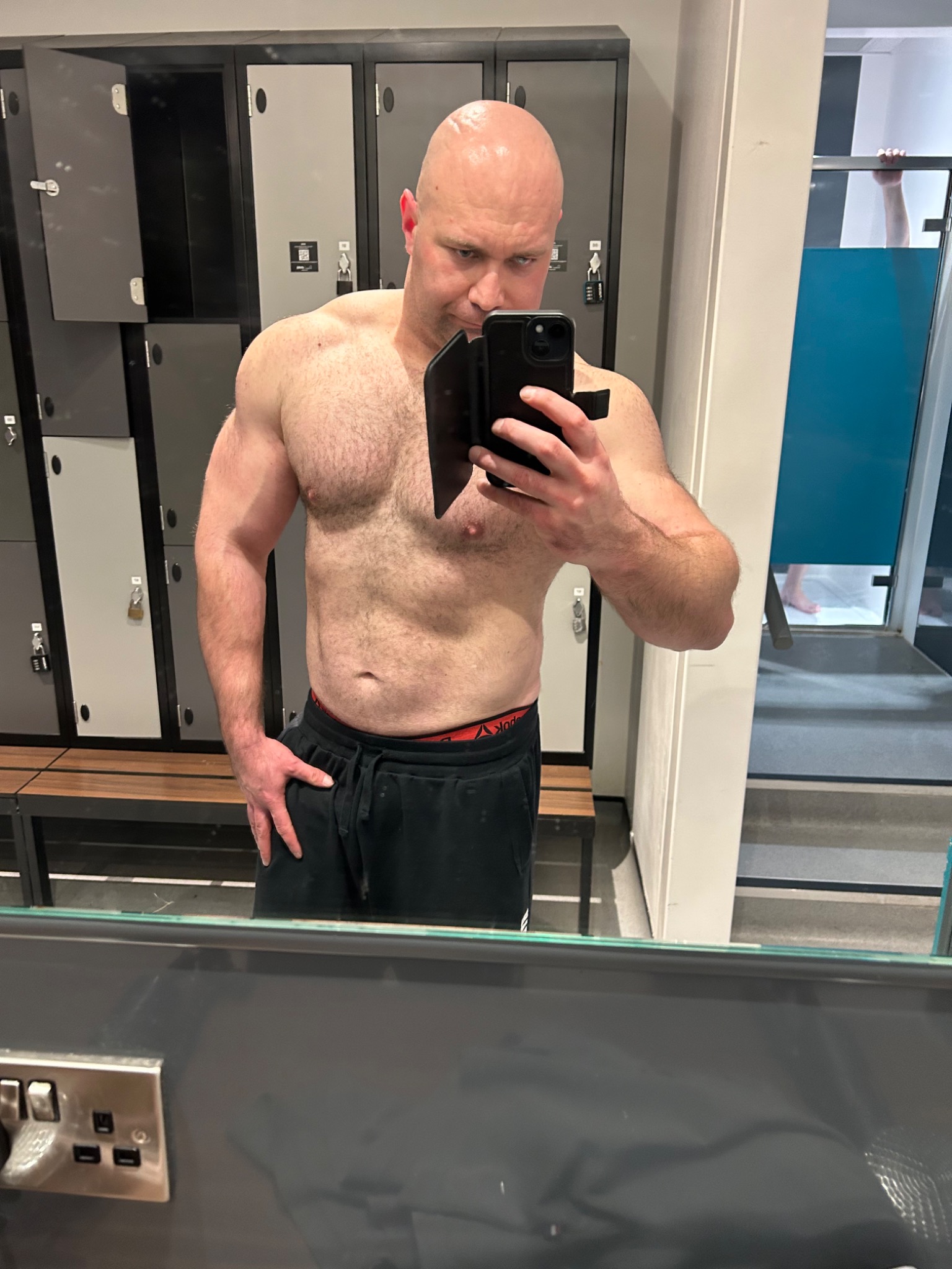 Mężczyzna bez koszulki robi selfie w szatni siłowni, widoczne szafki i ławka w tle.