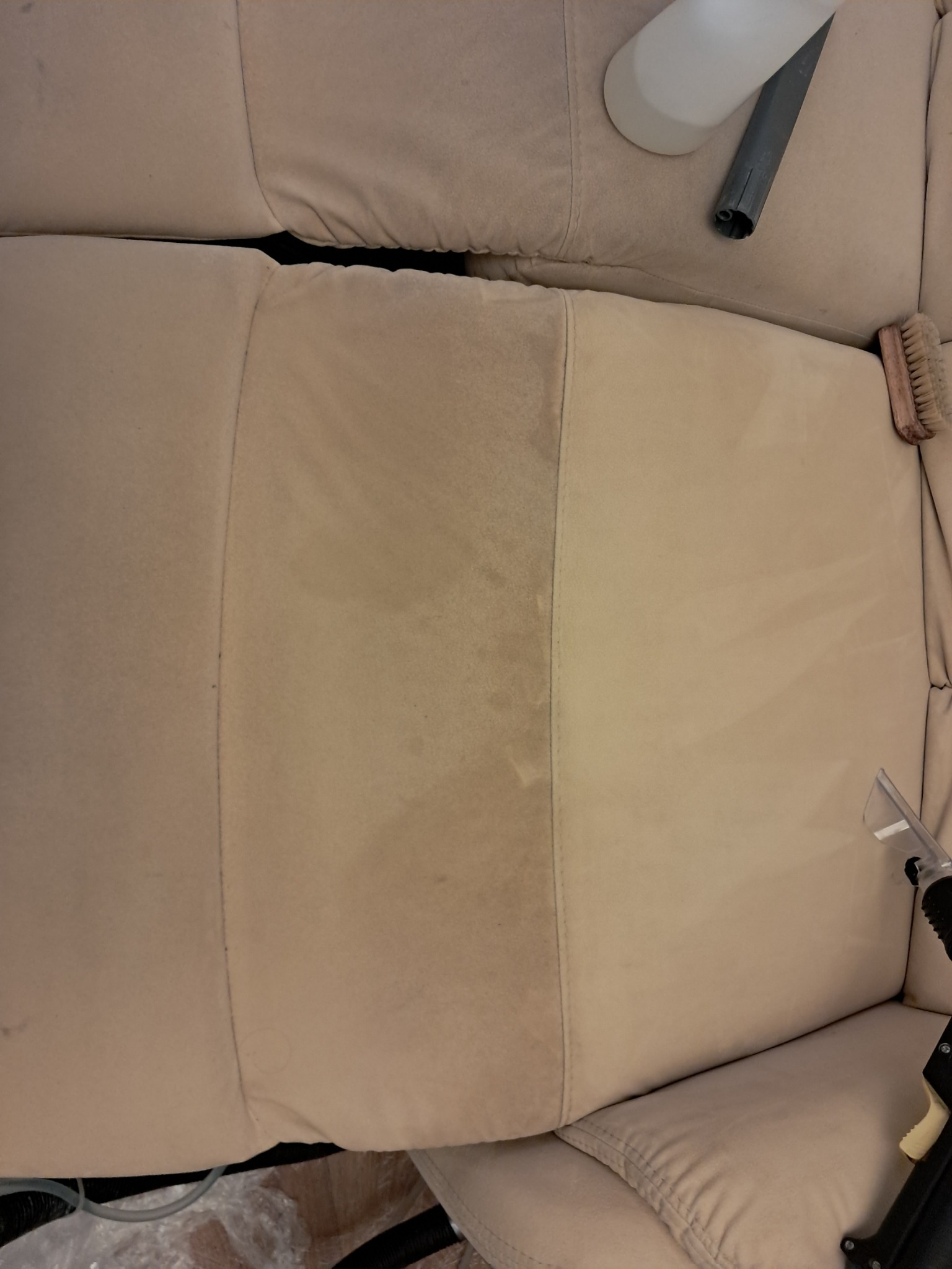 Fragment beżowej kanapy z widoczną różnicą czystości po czyszczeniu, narzędzia do czyszczenia tapicerki w tle.