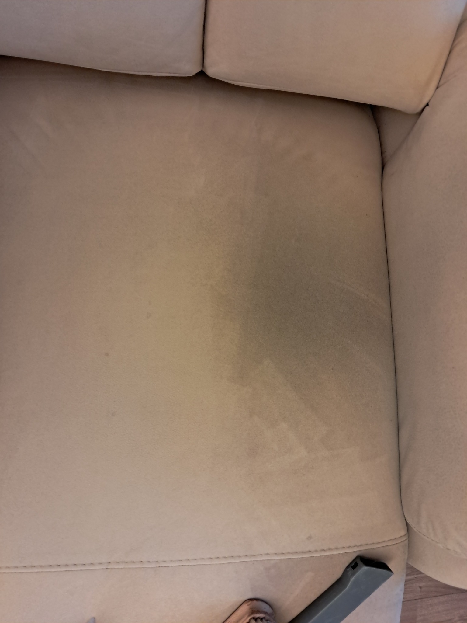 Jasna kanapa z wyraźnym, ciemnym zabrudzeniem, częściowo wyczyszczona z widoczną końcówką odkurzacza piorącego.