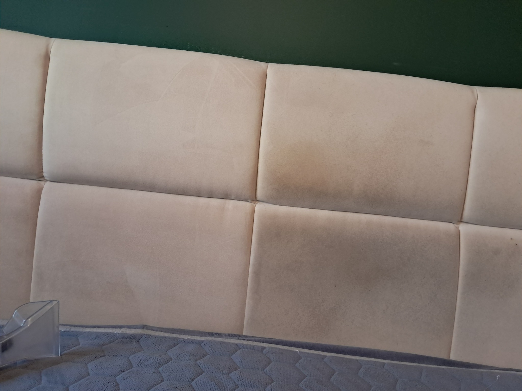 Beżowy zagłówek łóżka podzielony na kwadratowe panele, widoczne ślady użytkowania i plamy, fragment niebieskiego materaca na dole, dysza odkurzacza piorącego w lewym dolnym rogu.