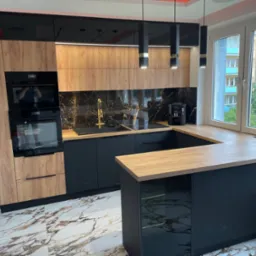 Nowoczesna kuchnia z czarnymi szafkami dolnymi i drewnopodobnymi górnymi, czarny marmurowy backsplash, złota armatura, wyspa z drewnianym blatem, marmurowa podłoga, widok z okna na budynek mieszkalny.