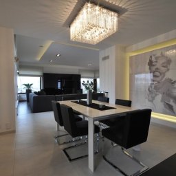 POLSKA FABRYKA MEBLI - Nowoczesna jadalnia z białym stołem, czarnymi krzesłami na chromowanych nogach, kryształowym żyrandolem i portretem Marilyn Monroe podświetlonym ledami.