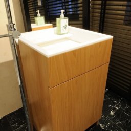 POLSKA FABRYKA MEBLI - Nowoczesna umywalka z szafką z jasnego drewna, chromowana bateria stojąca i czarny marmur na podłodze, odbicie w lustrze z żaluzjami.