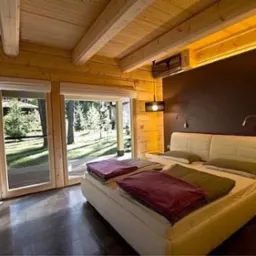 Drewniana sypialnia z dużym oknem na las, białe łóżko z kolorowymi pledami, drewniany sufit z belkami i oświetleniem punktowym, ciemna ściana za łóżkiem.