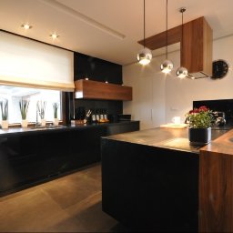 POLSKA FABRYKA MEBLI - Nowoczesna kuchnia z czarnymi, lakierowanymi frontami, drewnianym okapem i wyspą, oświetlona trzema chromowanymi lampami wiszącymi.