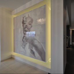 POLSKA FABRYKA MEBLI - Nowoczesny przedpokój z podświetlaną ścianą, na której znajduje się duży obraz Marilyn Monroe, widoczny fragment salonu z czarnymi meblami i korytarz z ciemną podłogą.