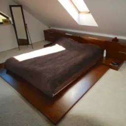 Minimalistyczna sypialnia na poddaszu z drewnianym łóżkiem na platformie, dopasowanymi szafkami nocnymi i oknem dachowym wpuszczającym jasne światło.