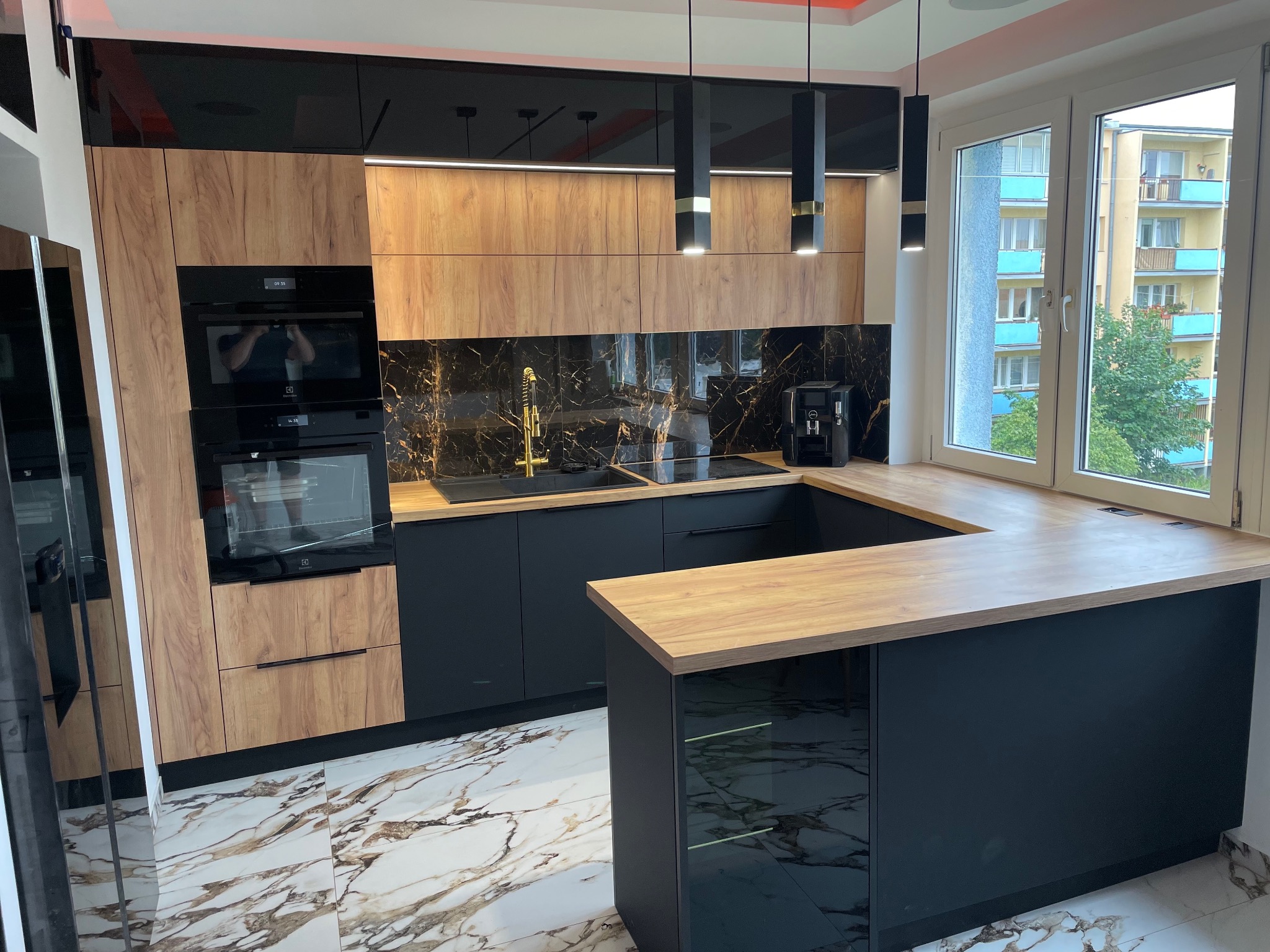 Nowoczesna kuchnia z czarnymi szafkami dolnymi i drewnopodobnymi górnymi, czarny marmurowy backsplash, złota armatura, wyspa z drewnianym blatem, marmurowa podłoga, widok z okna na budynek mieszkalny.