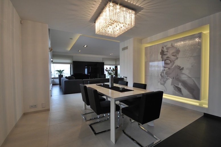 Nowoczesna jadalnia z białym stołem, czarnymi krzesłami na chromowanych nogach, kryształowym żyrandolem i portretem Marilyn Monroe podświetlonym ledami.