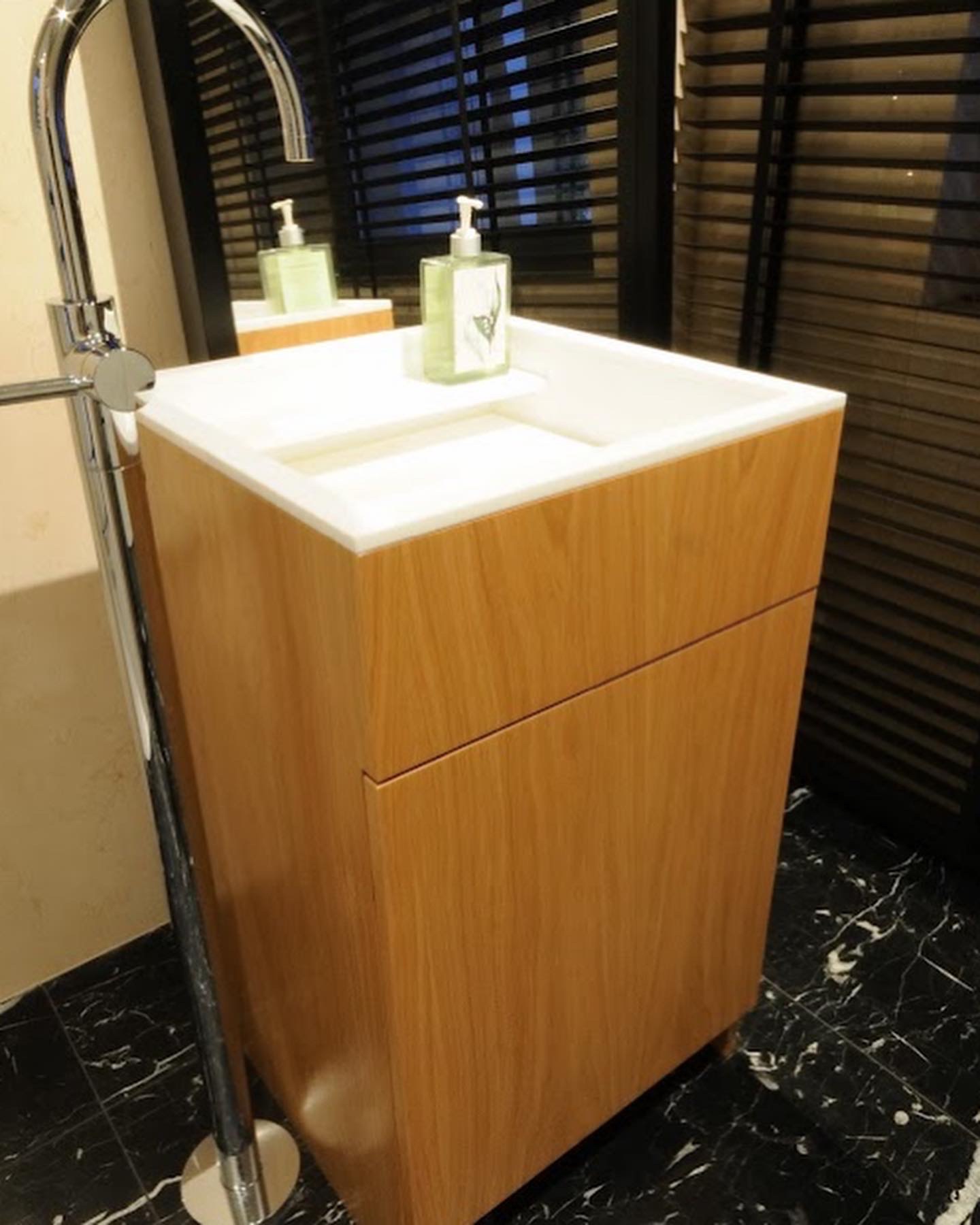 Nowoczesna umywalka z szafką z jasnego drewna, chromowana bateria stojąca i czarny marmur na podłodze, odbicie w lustrze z żaluzjami.