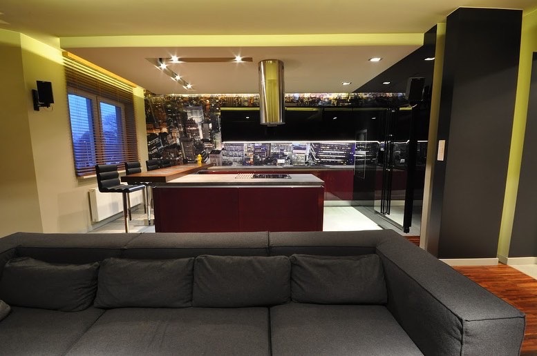 Nowoczesna kuchnia z wyspą w odcieniach czerwieni i czerni, fototapeta z panoramą miasta, widok z salonu z szarą sofą.