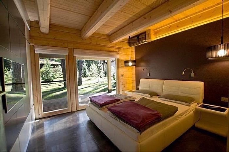 Drewniana sypialnia z dużym oknem na las, białe łóżko z kolorowymi pledami, drewniany sufit z belkami i oświetleniem punktowym, ciemna ściana za łóżkiem.