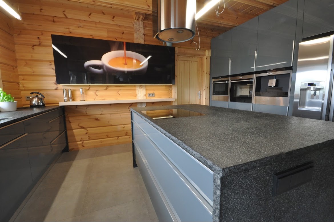 Nowoczesna kuchnia z drewnianymi ścianami, szarymi szafkami i wyspą z blatem z ciemnego granitu, nad którą wisi okap; na ścianie duży obraz z motywem kawy.