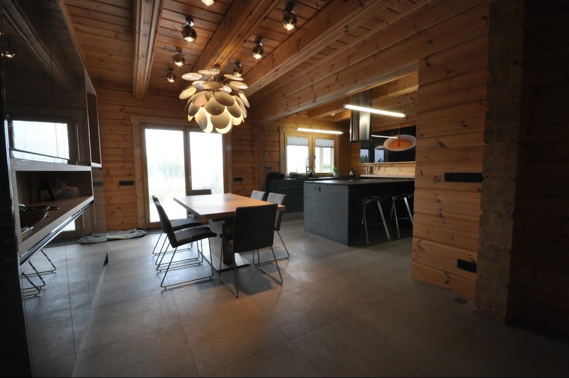 Wnętrze domu z drewnianymi ścianami, widoczna nowoczesna kuchnia z wyspą, stół z krzesłami i designerska lampa sufitowa.