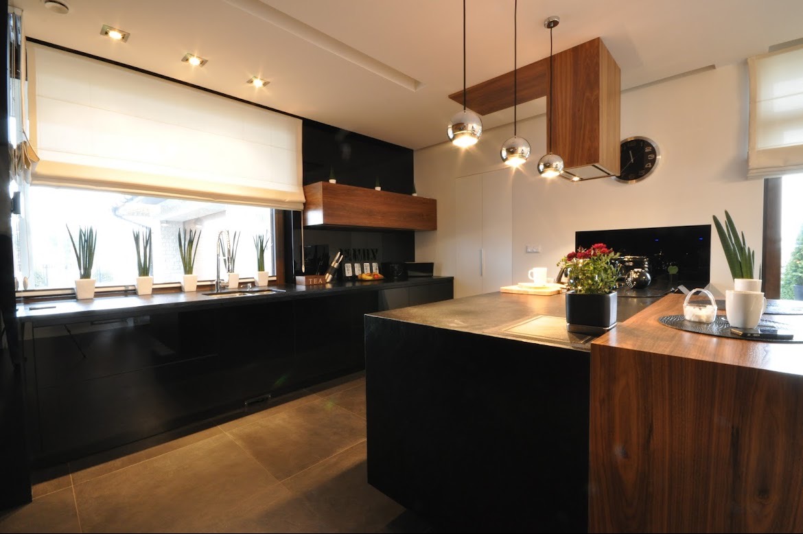 Nowoczesna kuchnia z czarnymi, lakierowanymi frontami, drewnianym okapem i wyspą, oświetlona trzema chromowanymi lampami wiszącymi.