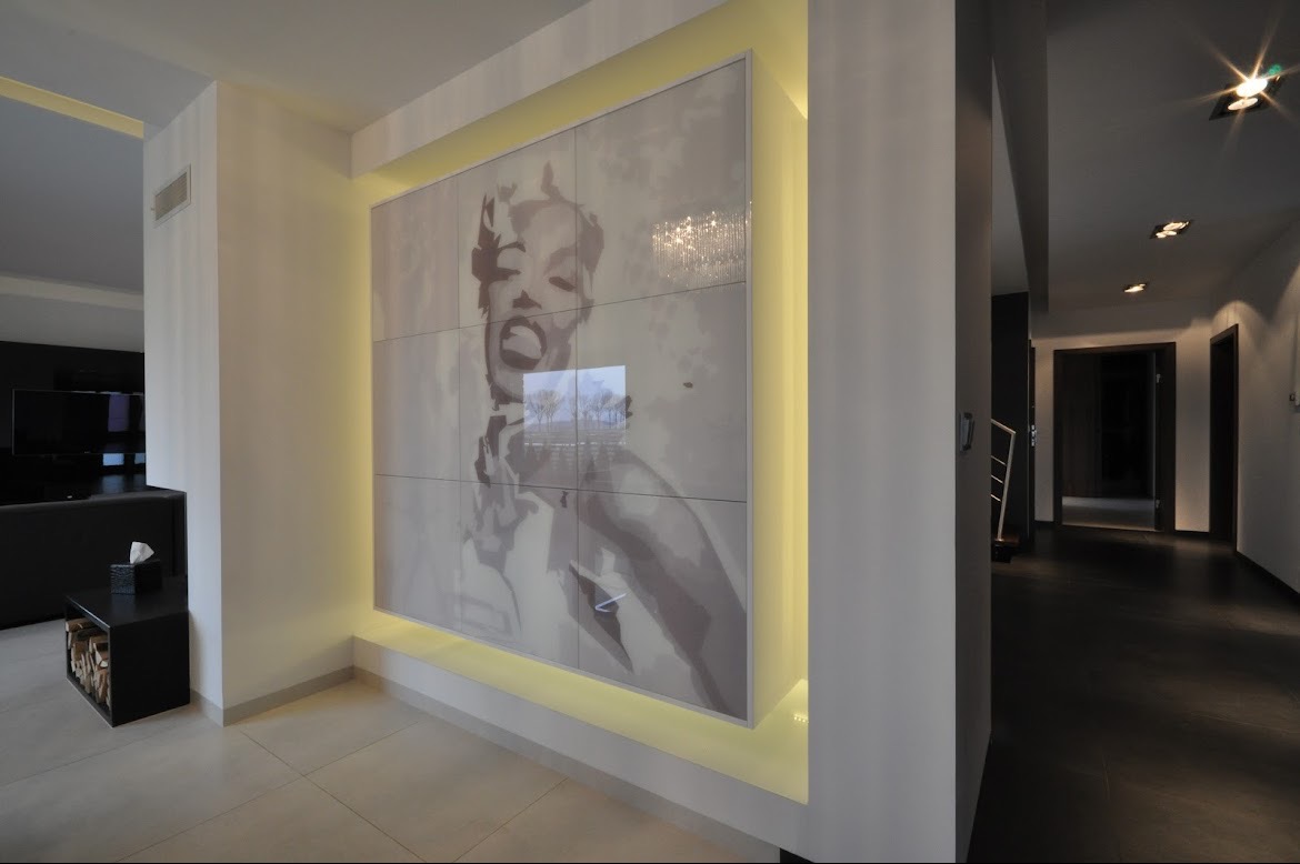 Nowoczesny przedpokój z podświetlaną ścianą, na której znajduje się duży obraz Marilyn Monroe, widoczny fragment salonu z czarnymi meblami i korytarz z ciemną podłogą.